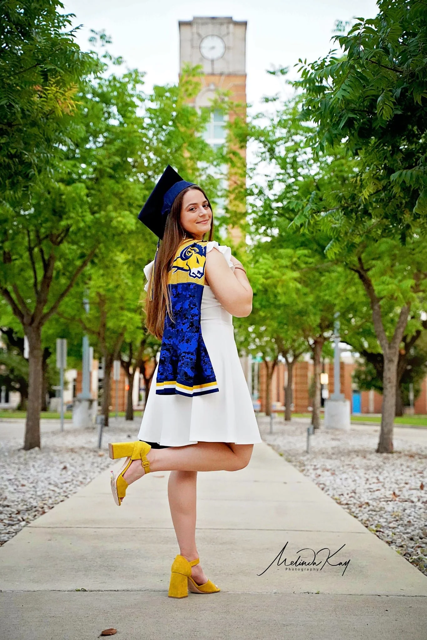 Young woman in a white dress and yellow high heels, wearing a navy blue graduate cap and a blue and gold stole, standing on a sidewalk in a park with green trees, with a clock tower in the background.