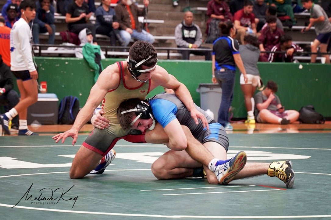 Two wrestlers competing on a mat, with one in a red and beige singlet and the other in a blue and black singlet, engaged in a takedown move. Spectators are visible in the background.