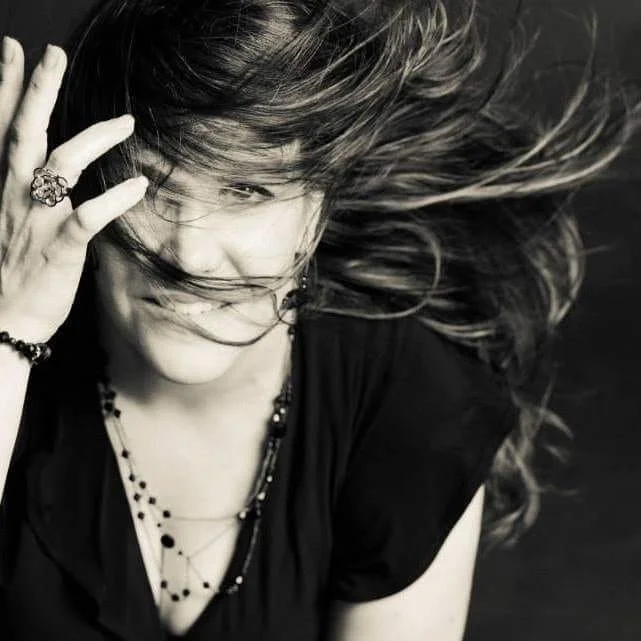 Black and white photo of a woman with long hair holding her head and smiling slightly, looking at the camera, wearing layered necklaces and a ring.