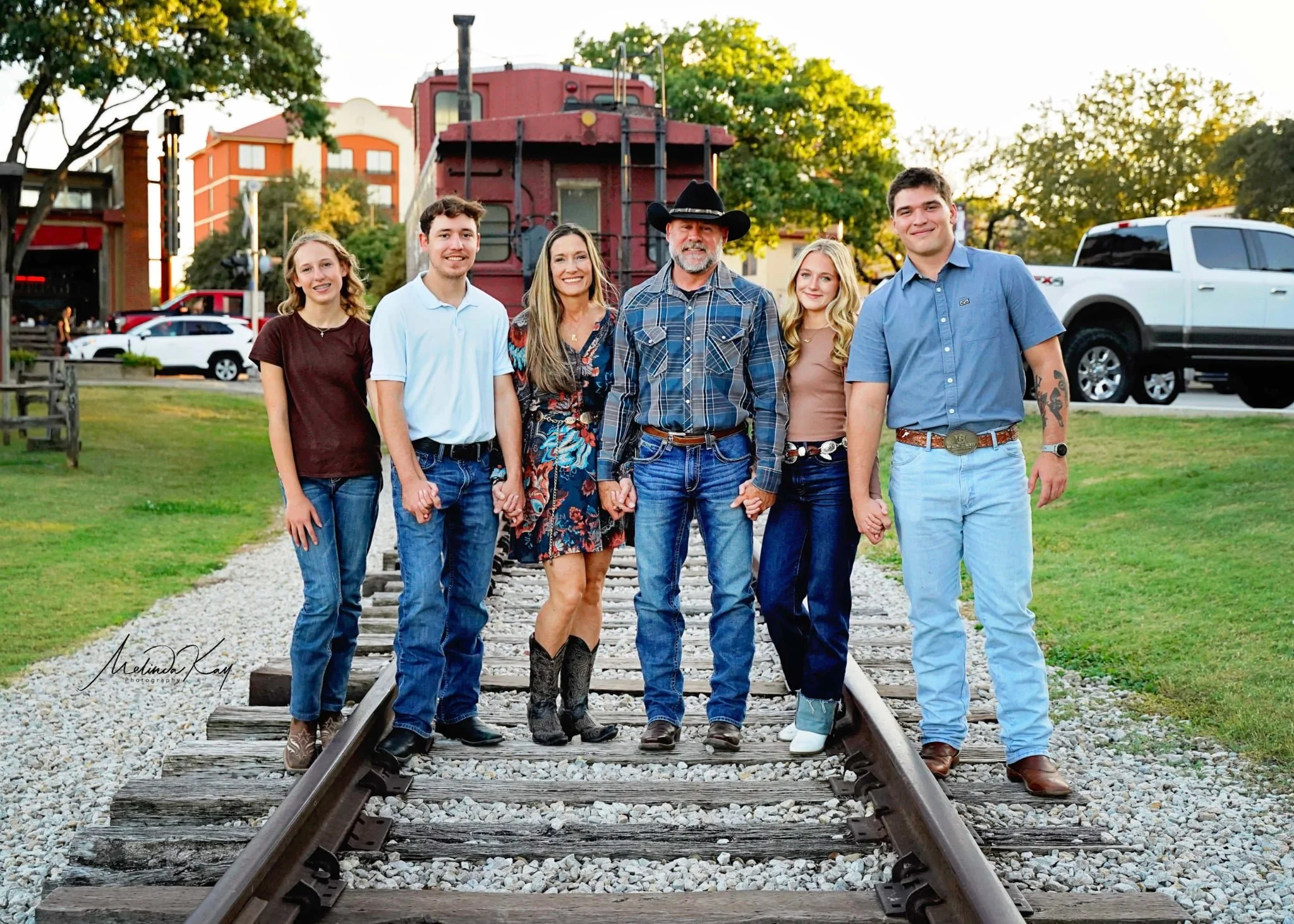 Group of six people standing on train tracks holding hands outside in park, smiling.