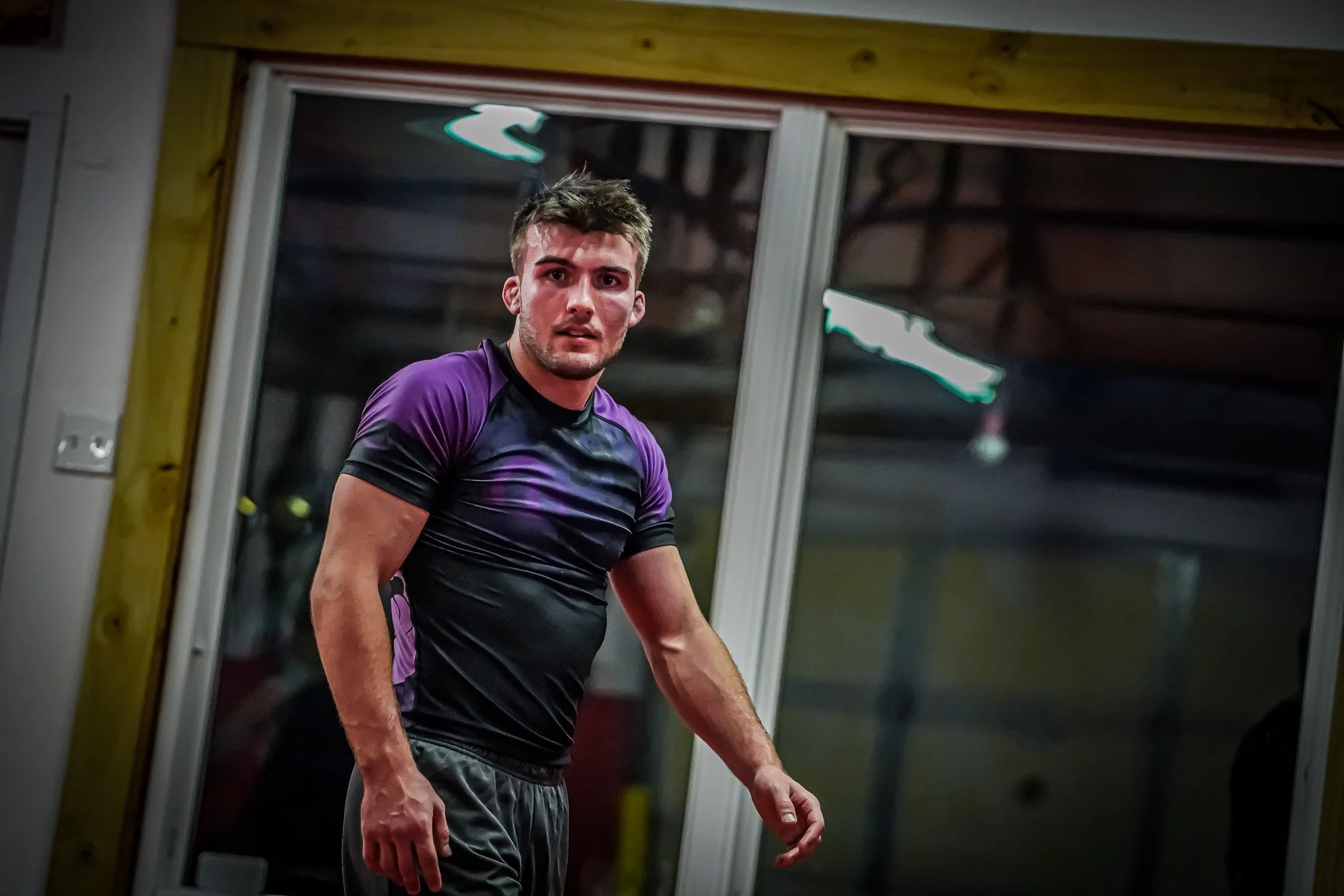A young man with short, slightly messy hair, wearing a purple and black athletic shirt and black shorts, standing indoors near large glass windows, looking towards the camera with a serious expression.