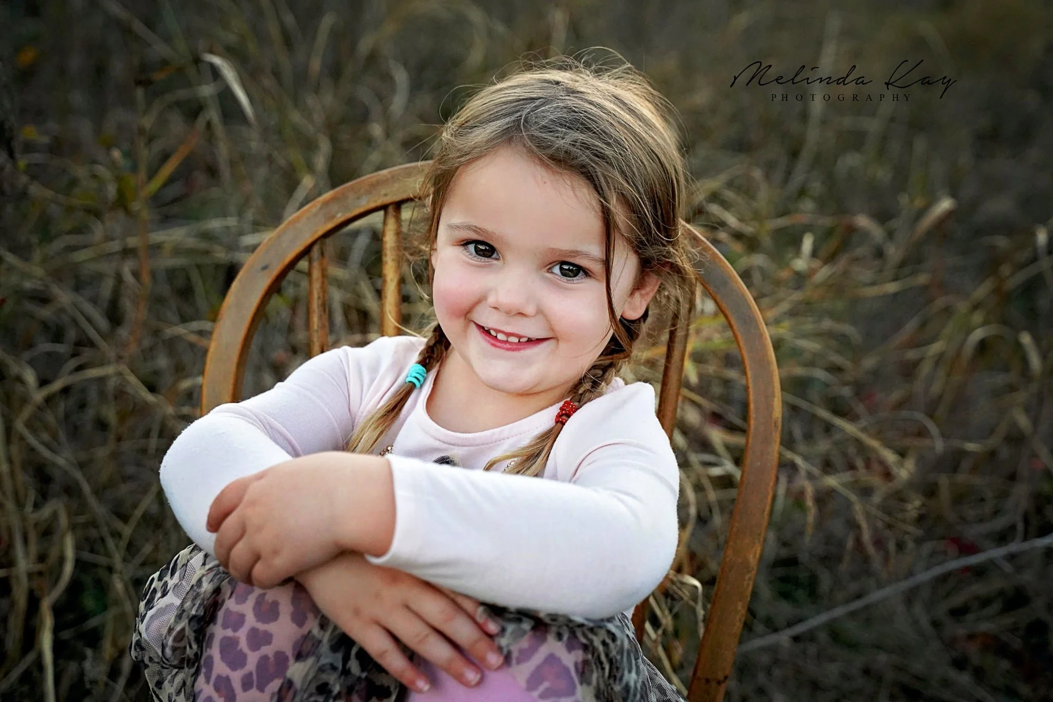 A young girl with braided hair, smiling and sitting on a rusty metal chair outdoors, surrounded by dried reeds or grasses.
