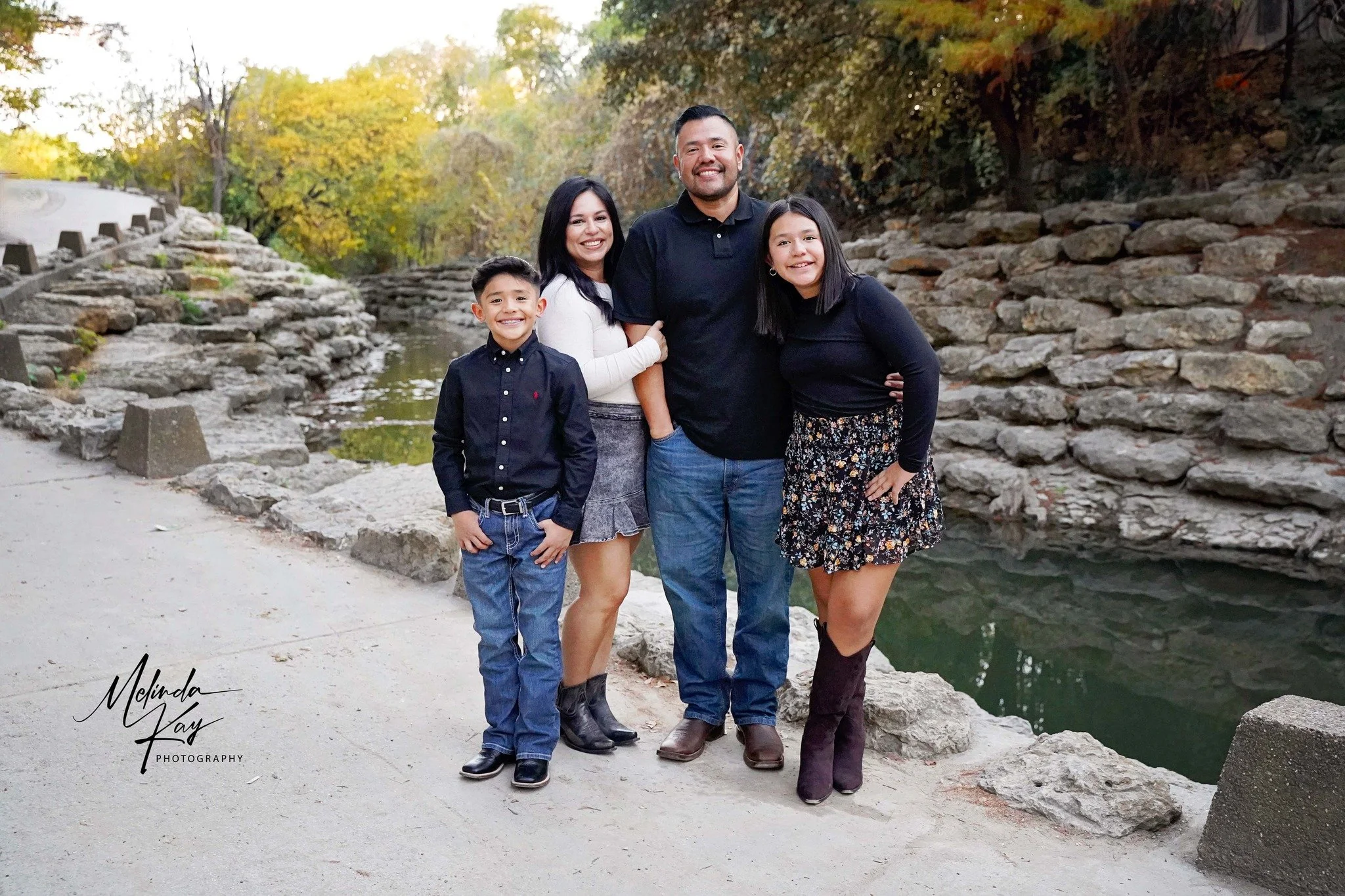 Family of four standing outdoors by a creek with rocks and autumn trees in the background, smiling at the camera.