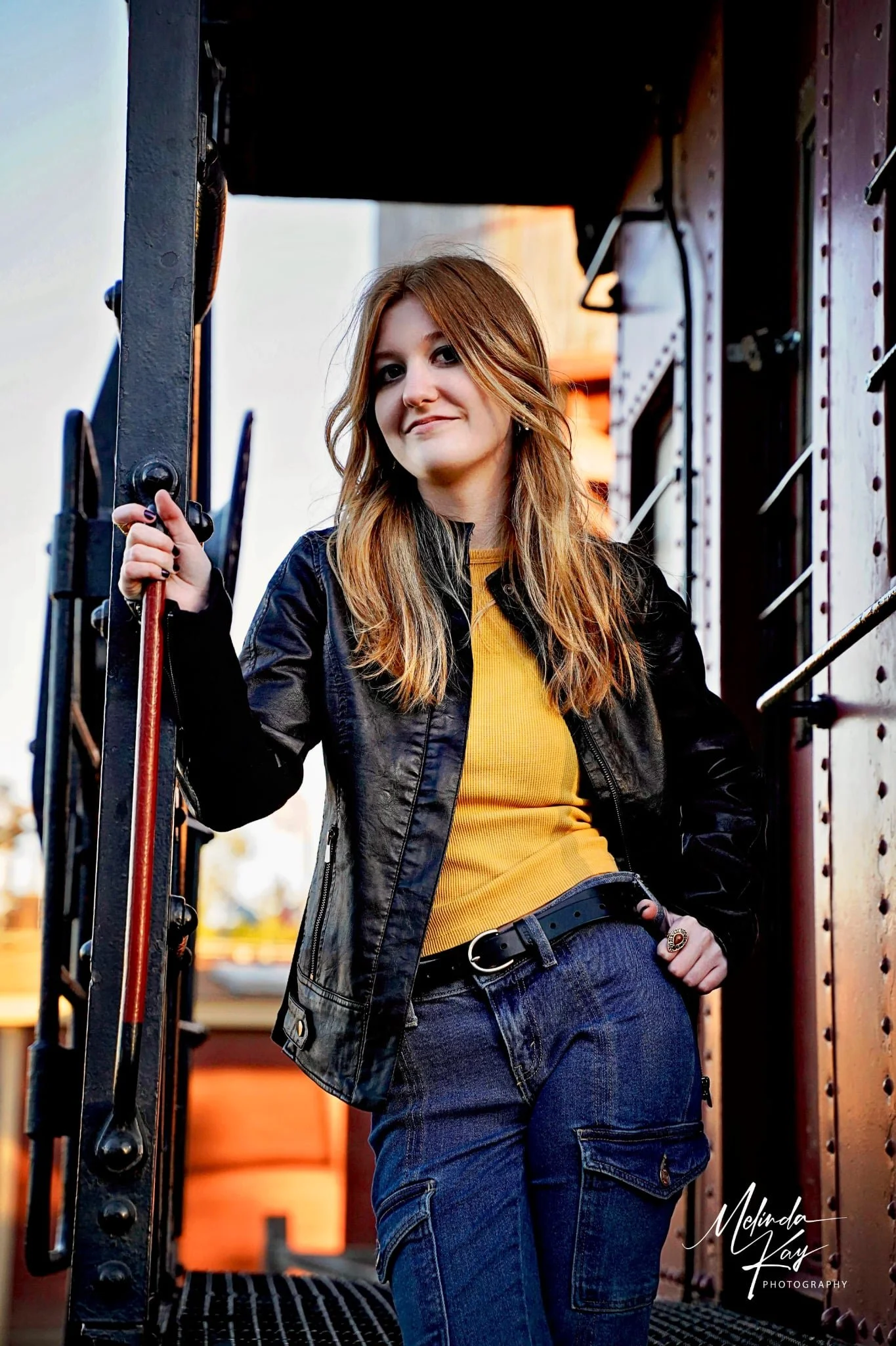 A young woman with long, wavy red hair stands on a train or train platform, wearing a black leather jacket, yellow shirt, and blue jeans. She is holding onto a metal railing and looking at the camera with a confident expression.