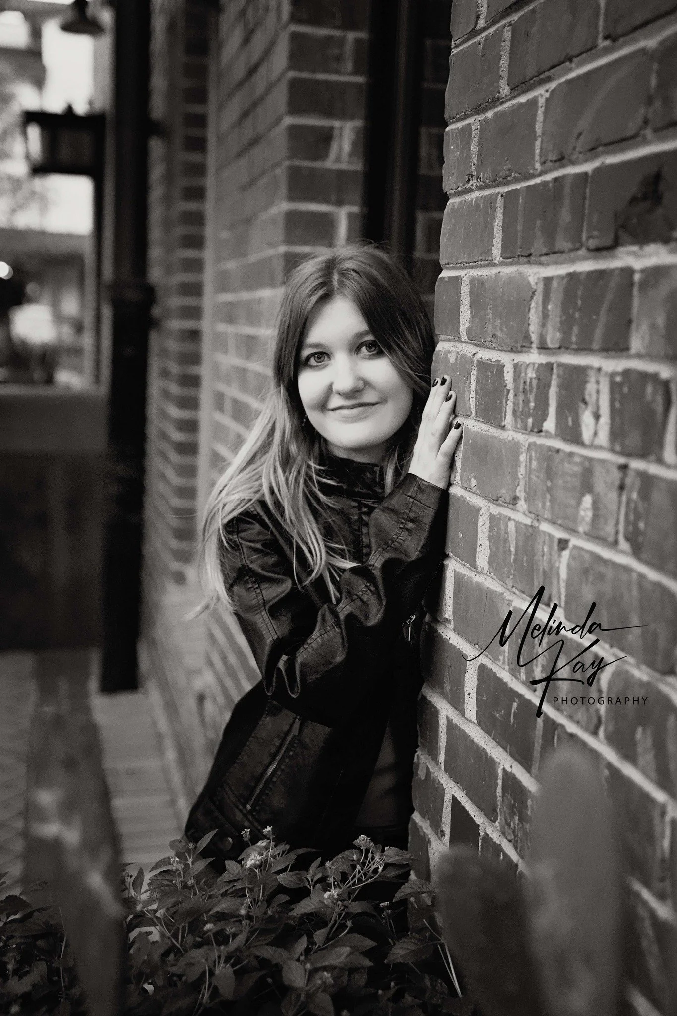 A woman with long hair wearing a leather jacket, standing beside a brick wall with her hand resting on it, smiling at the camera in black and white.
