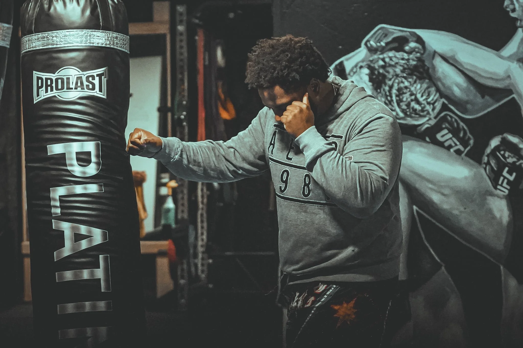 A person practicing martial arts in a gym, punching a black ProLast punching bag with white lettering, while wearing a gray hoodie and dark shorts.