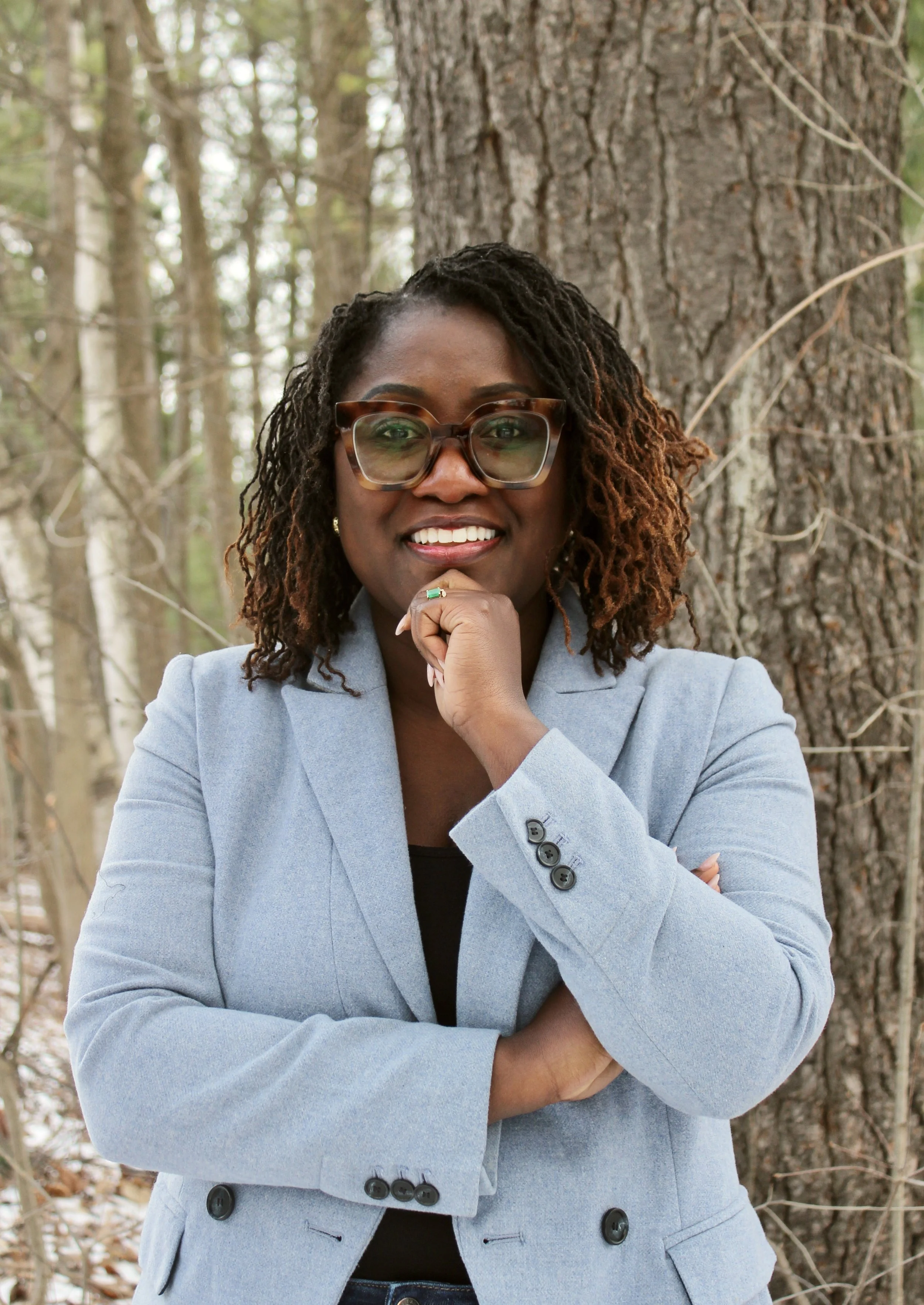 A woman wearing glasses and a light blue blazer, smiling with her hand under her chin, standing outdoors in front of a tree.