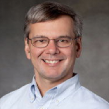 Portrait of a man with glasses and short gray hair, smiling, wearing a light blue collared shirt.