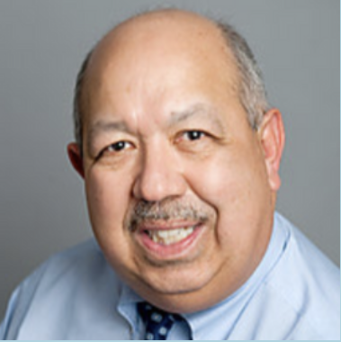 Close-up of a smiling middle-aged man with light skin, bald on top, wearing a light blue shirt and a patterned tie, against a gray background.