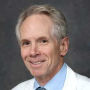 A portrait of a smiling middle-aged man with gray hair, wearing a light blue shirt and a white blazer against a dark gray background.