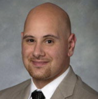Headshot of a man with a bald head, light skin, and facial hair, wearing a beige suit and tie, smiling against a neutral background.