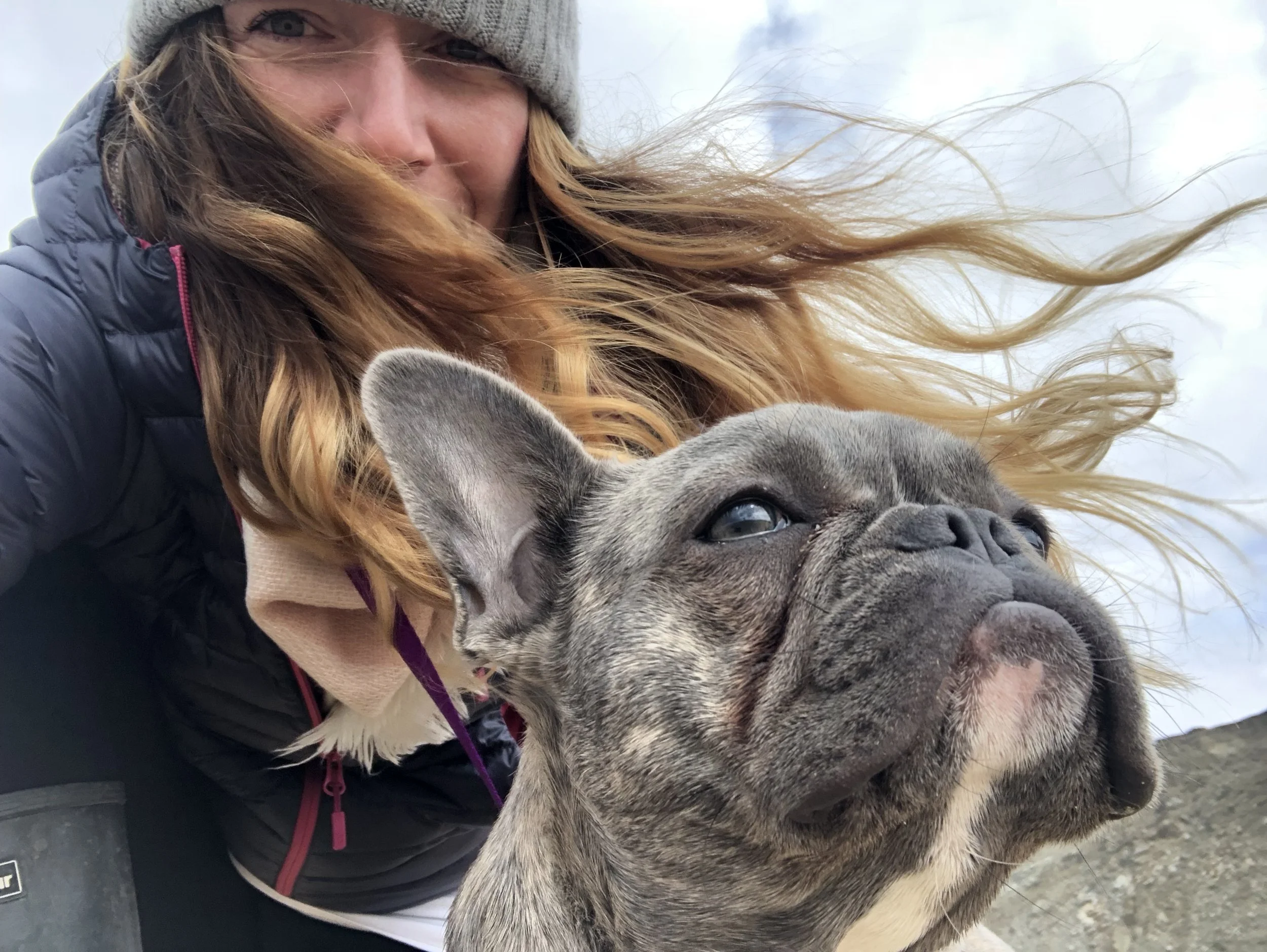 Beth with her long, wavy hair blowing across her face whilst holding Mabel her French Bulldog.