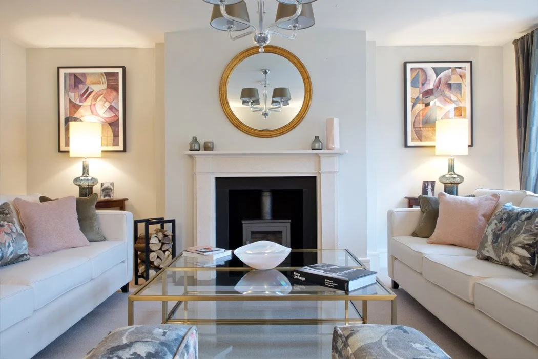 Living room with two white sofas, decorative pillows, a glass coffee table, a fireplace with a round mirror above, wall art, table lamps, and a ceiling chandelier.