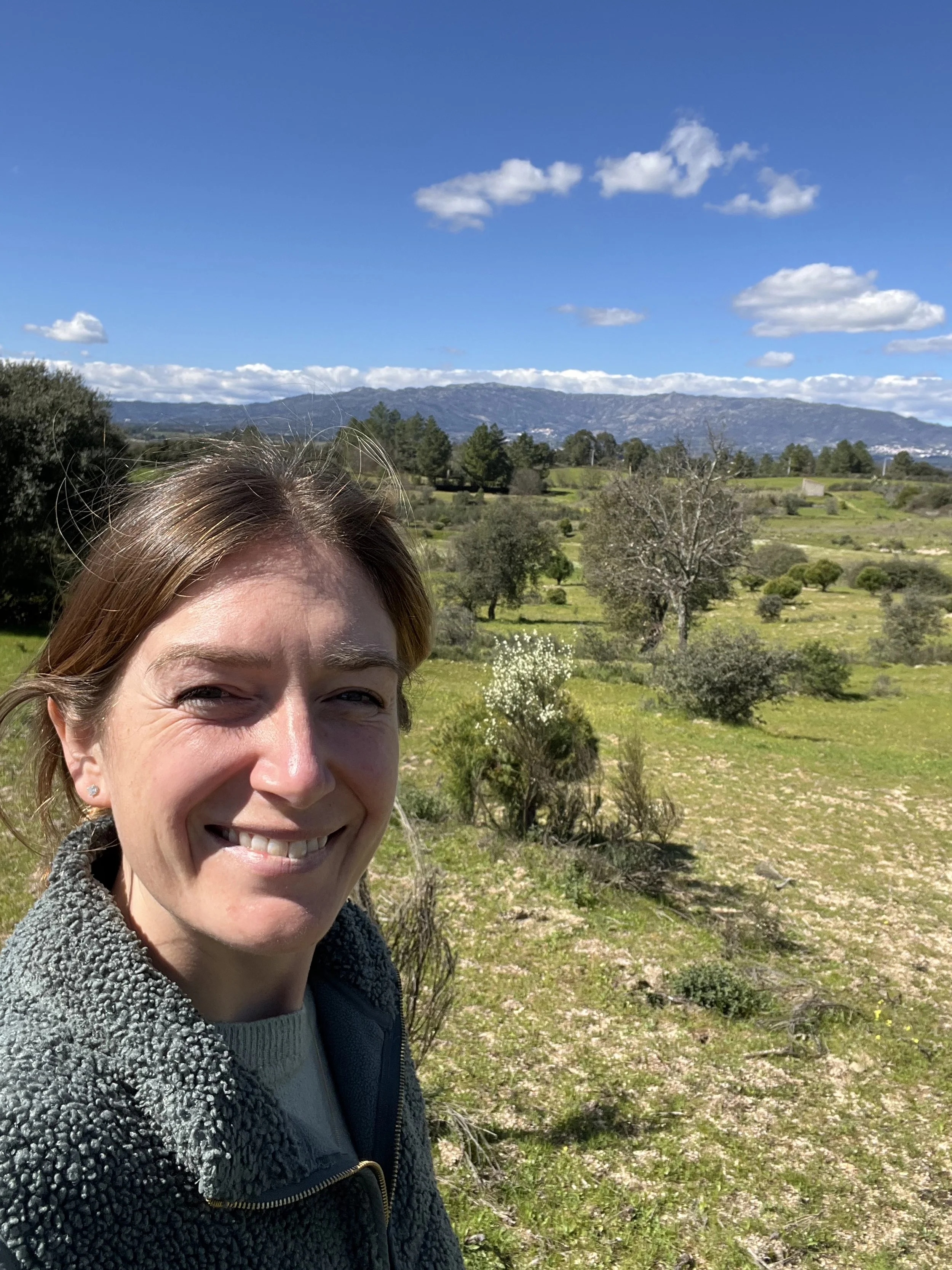 Beth smiling outdoors in a scenic green landscape with trees, mountains, and a blue sky with clouds in the background.