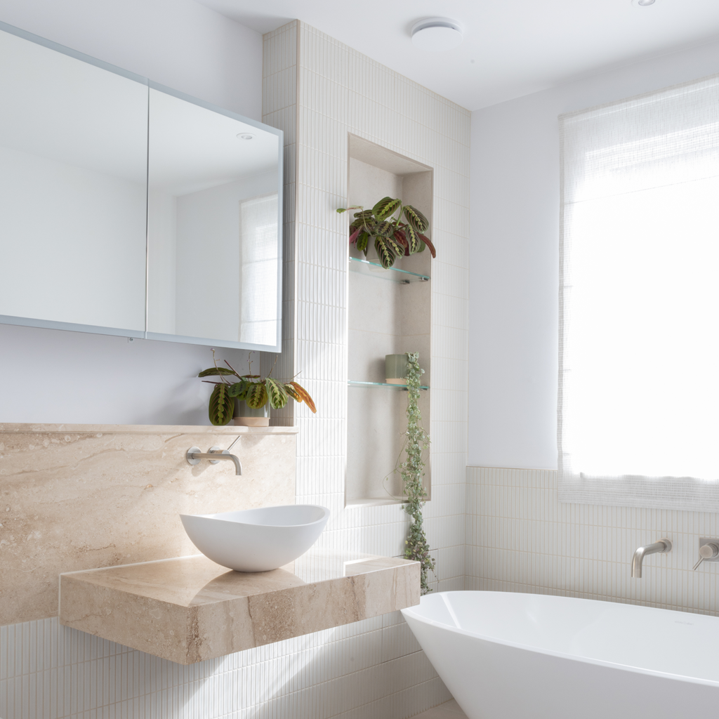 Bright bathroom featuring a freestanding bathtub, a floating beige marble vanity with a vessel sink, a large mirror, two potted plants, glass shelves with decorative items, and a window with a sheer curtain.