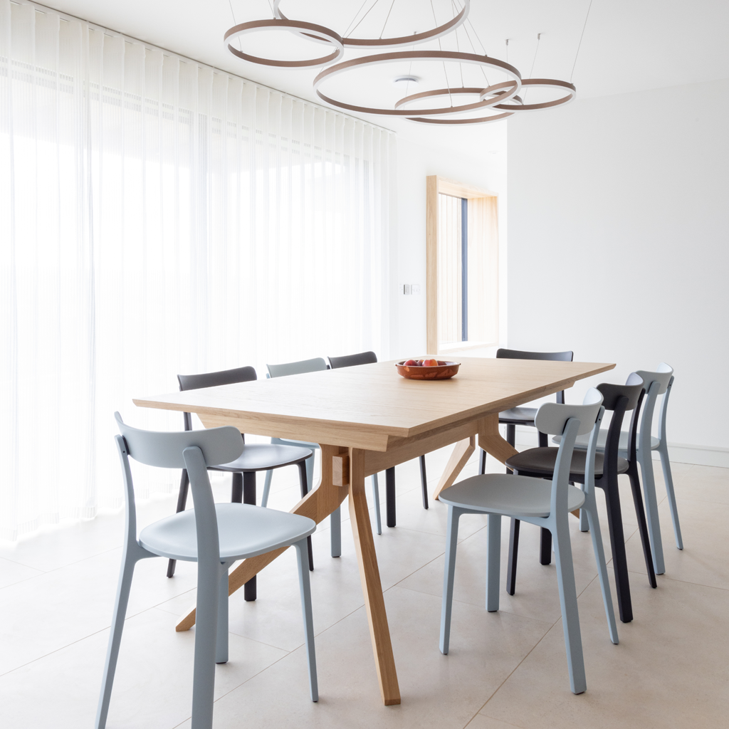 Modern dining room with a light wood table and eight chairs, three black and five white, in front of large glass windows with sheer curtains, and a unique circular light fixture hanging from the ceiling.