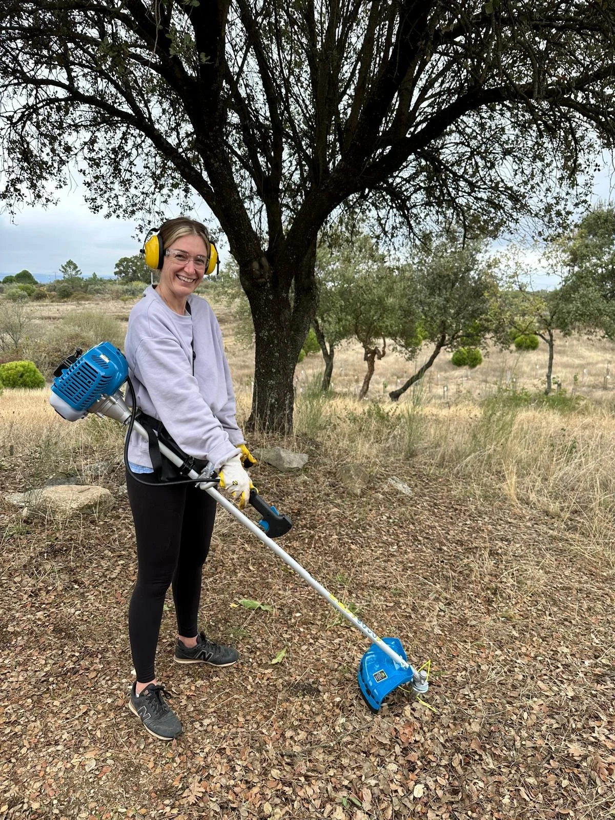 Beth wearing safety headphones and gloves is smiling while using a strimmer on her land in Portugal.