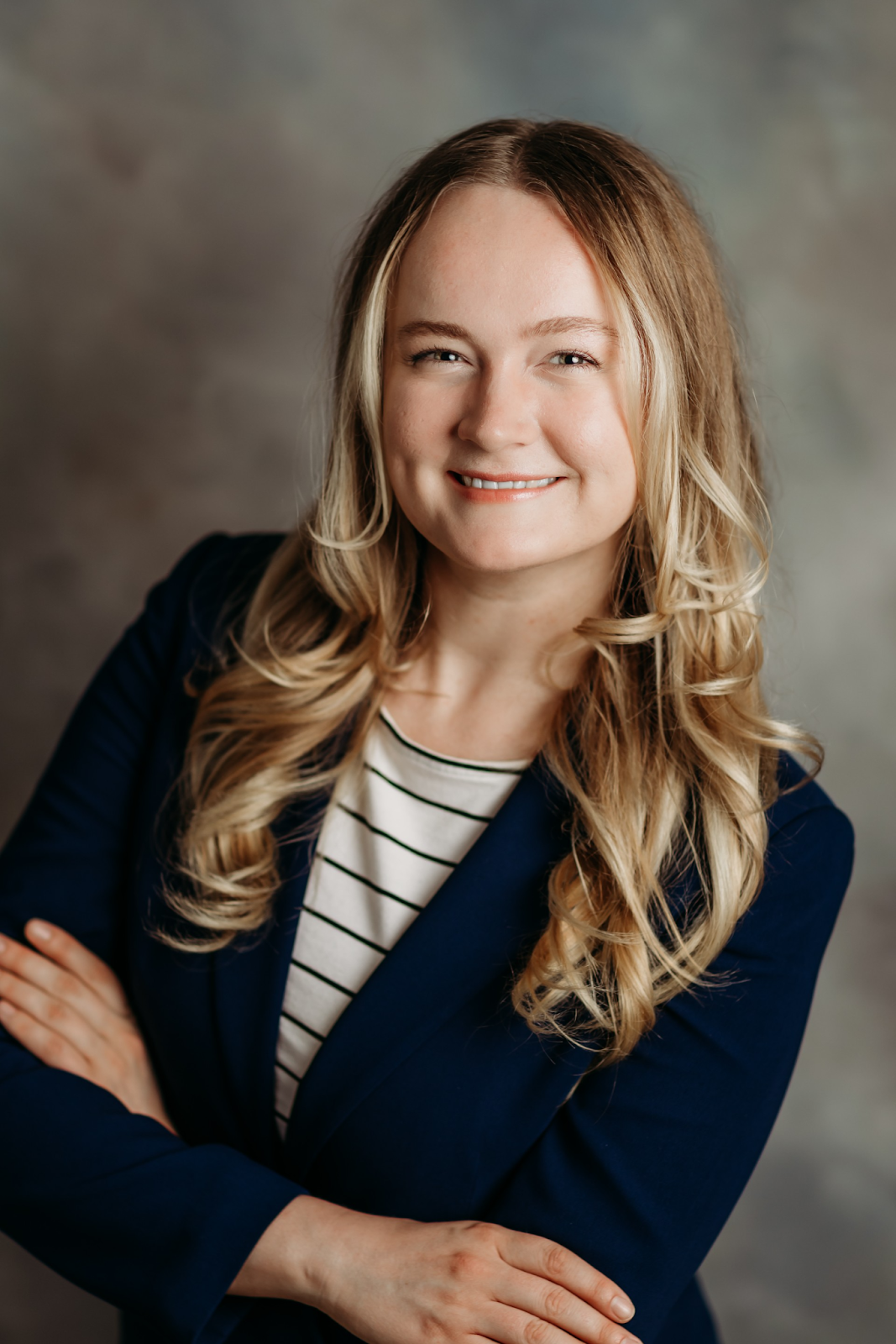 A professional woman with blonde hair, smiling, wearing a navy blazer and a striped shirt, with arms crossed in front of a neutral background.