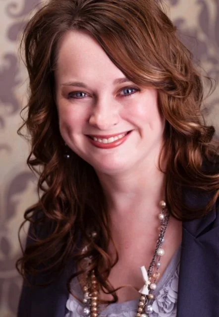 Portrait of a woman with long, curly brown hair, smiling, wearing a navy blazer, a light-colored blouse, and a pearl necklace.