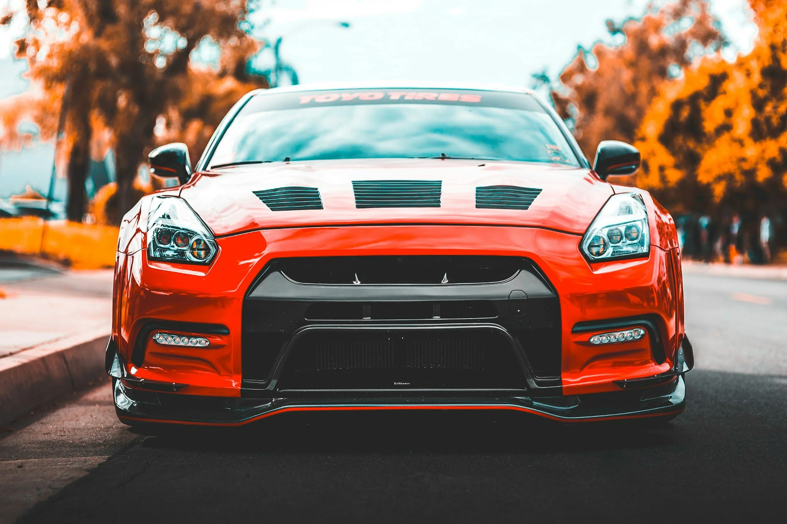 Front view of a red sports car with black accents parked on a street with trees in the background.