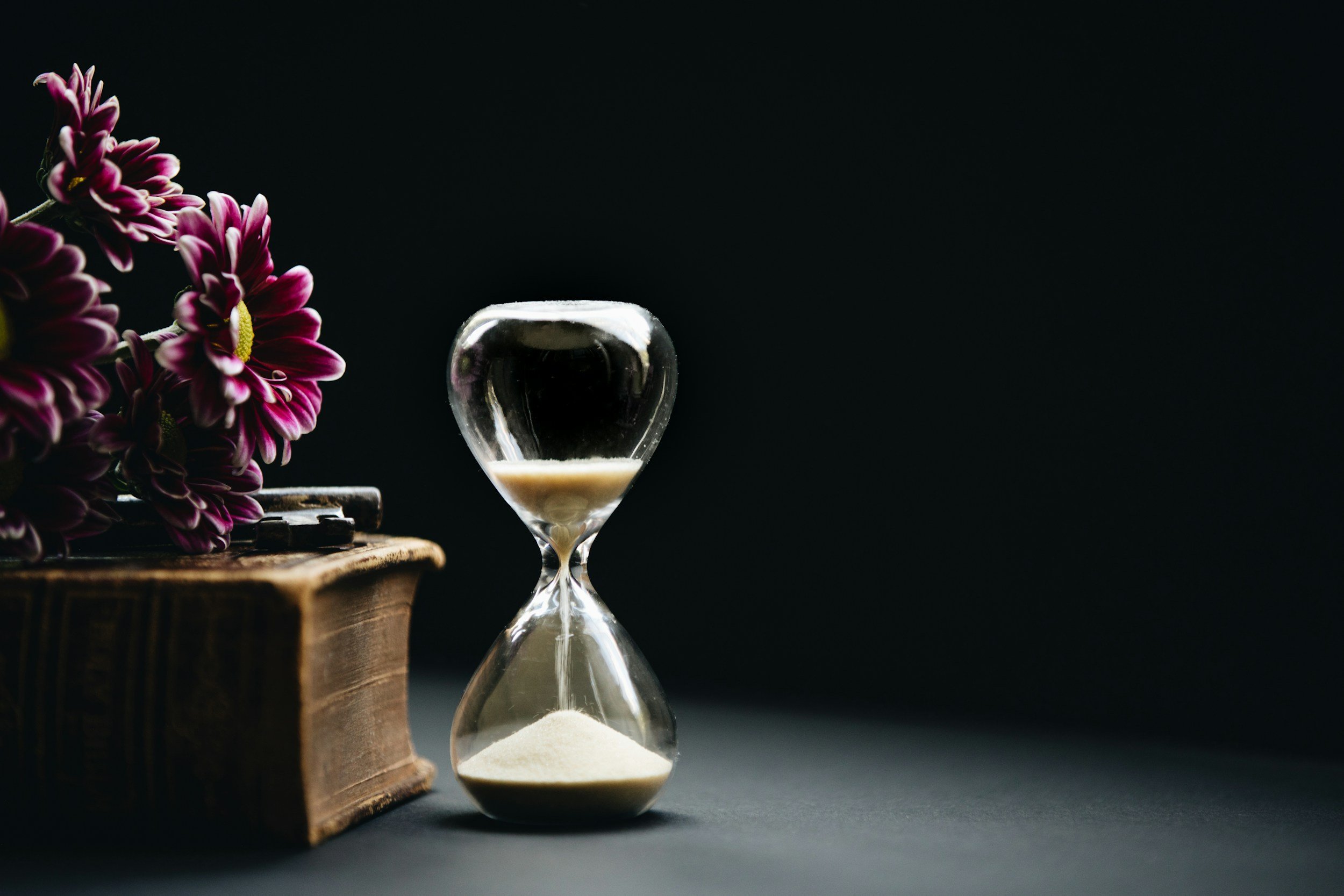 An hourglass with white sand placed on a dark surface next to a vintage book and a bouquet of purple and white daisy flowers, all against a black background.