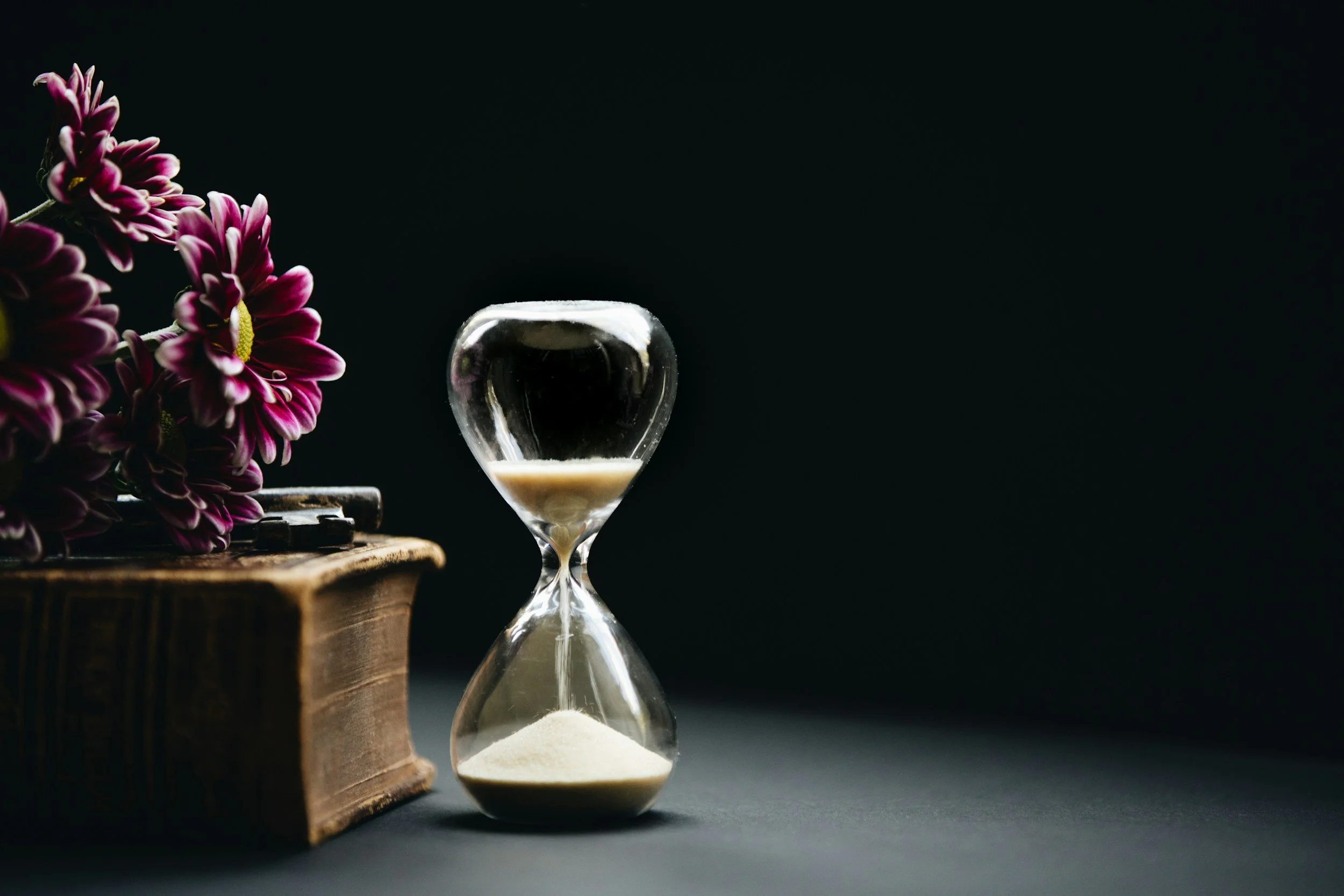 An hourglass with white sand, a vintage book, and purple flowers on a dark surface against a black background.