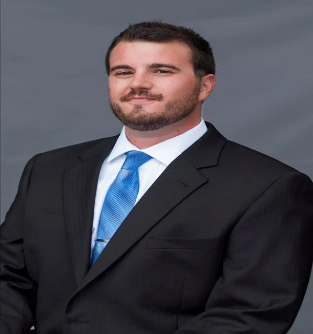 A man in a black suit with a light blue tie, standing against a plain gray background.