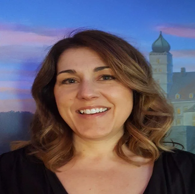 A woman with shoulder-length brown hair smiling in front of a painted background featuring a building with a dome and a tower.