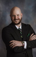 Professional man with a beard, wearing a suit and tie, smiling with arms crossed, against a gray background.