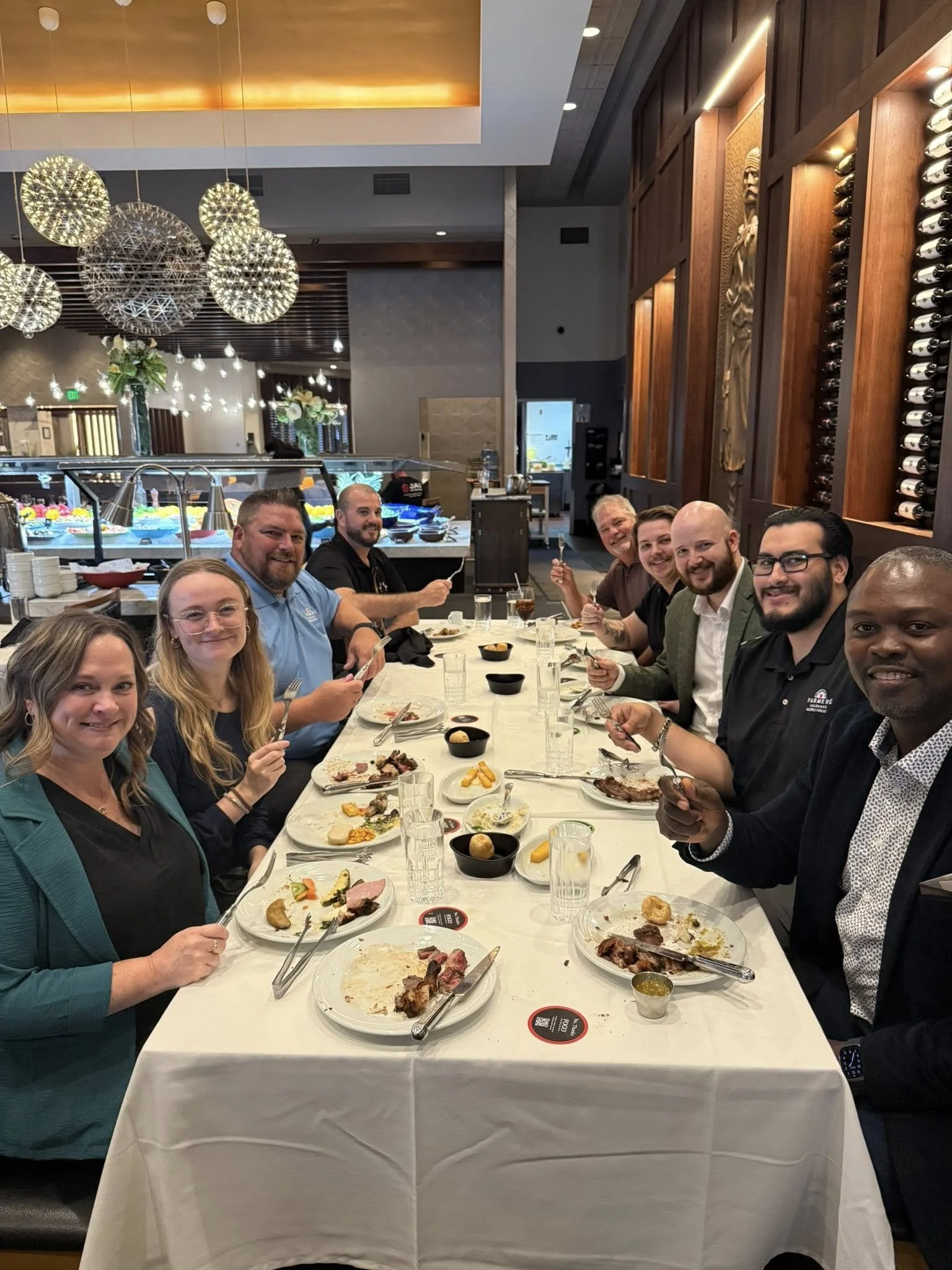 A group of nine people sitting at a long dinner table in a restaurant, smiling and enjoying their meal.