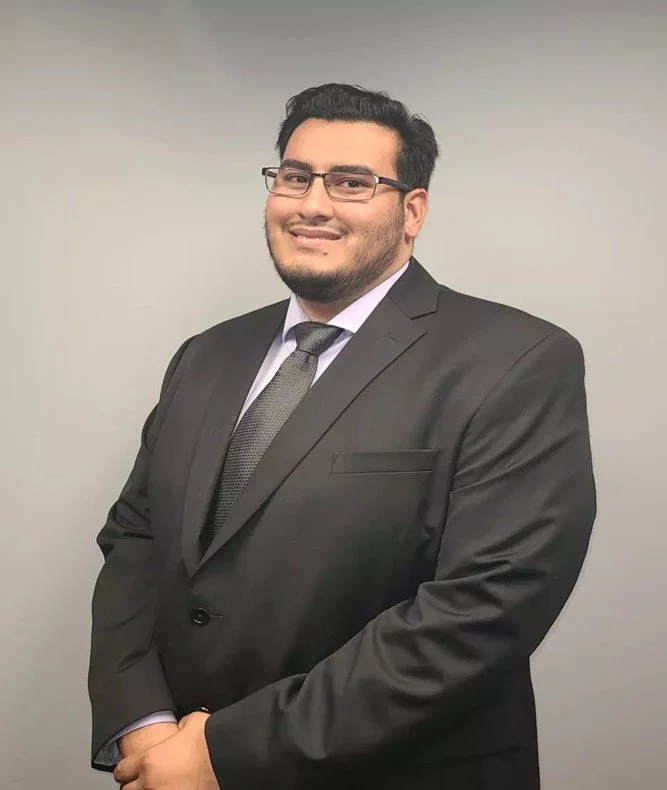 A man in a dark suit, white shirt, and patterned tie standing against a plain gray background, smiling at the camera.