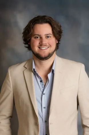Headshot of a young man with medium-length curly hair, smiling, wearing a beige blazer and blue dress shirt, against a blurred gray background.