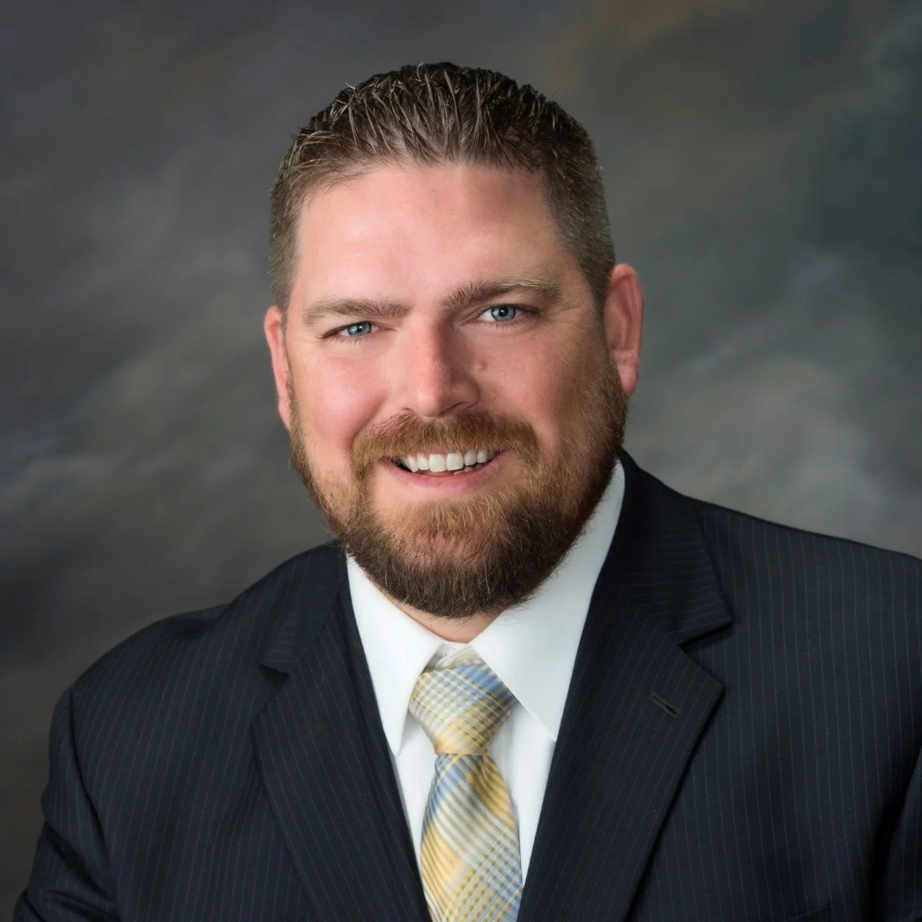 Headshot of a smiling man with a beard and blue eyes, wearing a dark pinstripe suit and a yellow striped tie, against a blurred dark background.
