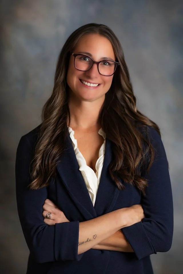 A woman with long brown hair, wearing glasses, a white blouse, and a navy blazer, smiling with arms crossed, against a neutral background.