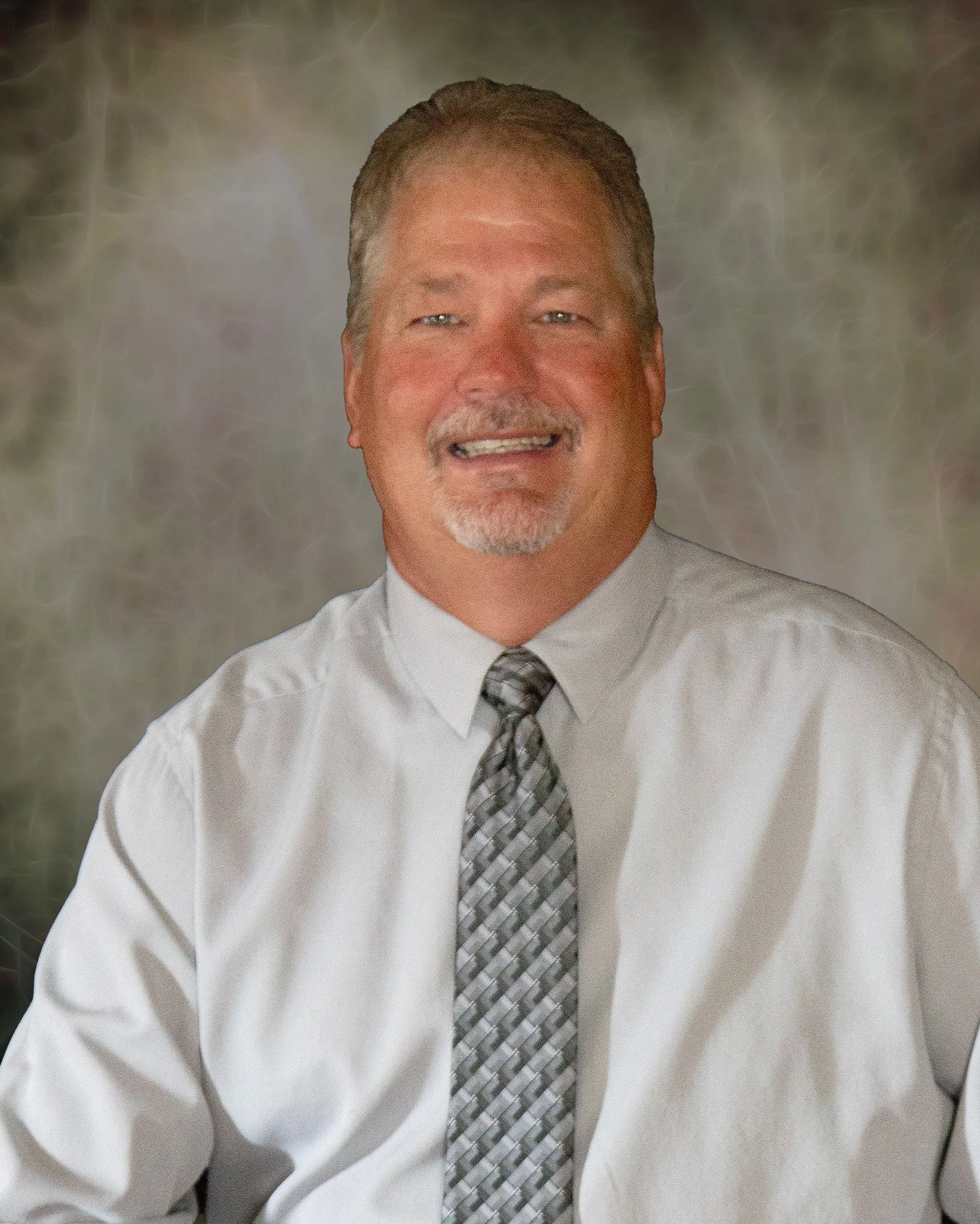 A middle-aged man with gray hair, a goatee, wearing a white dress shirt and a patterned gray tie, smiling at the camera.