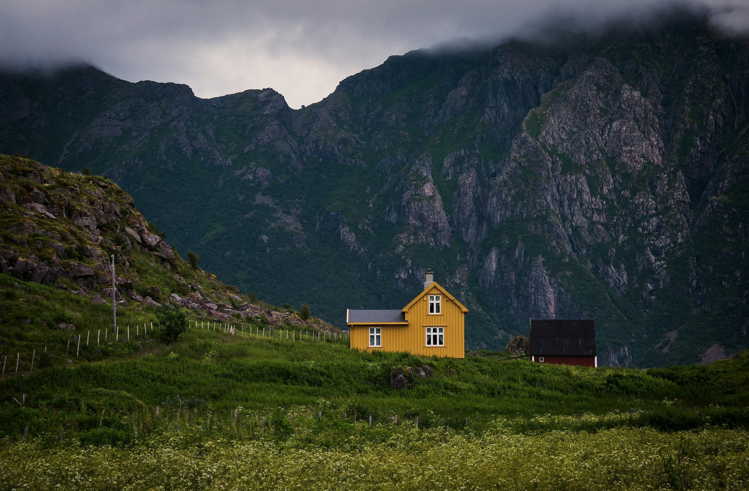 A yellow house with white windows on a green hill, with a mountain range in the background and cloudy sky above.