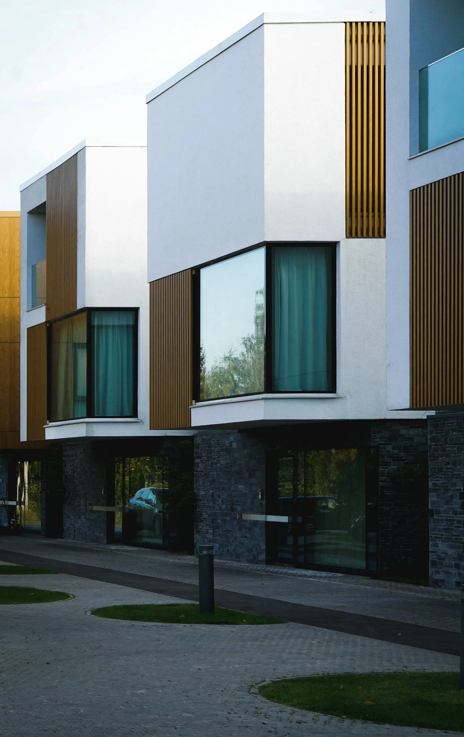 Modern residential building with white exterior walls, large windows with blue curtains, wooden accents, and stone foundation, with a sidewalk and small patches of grass in front.
