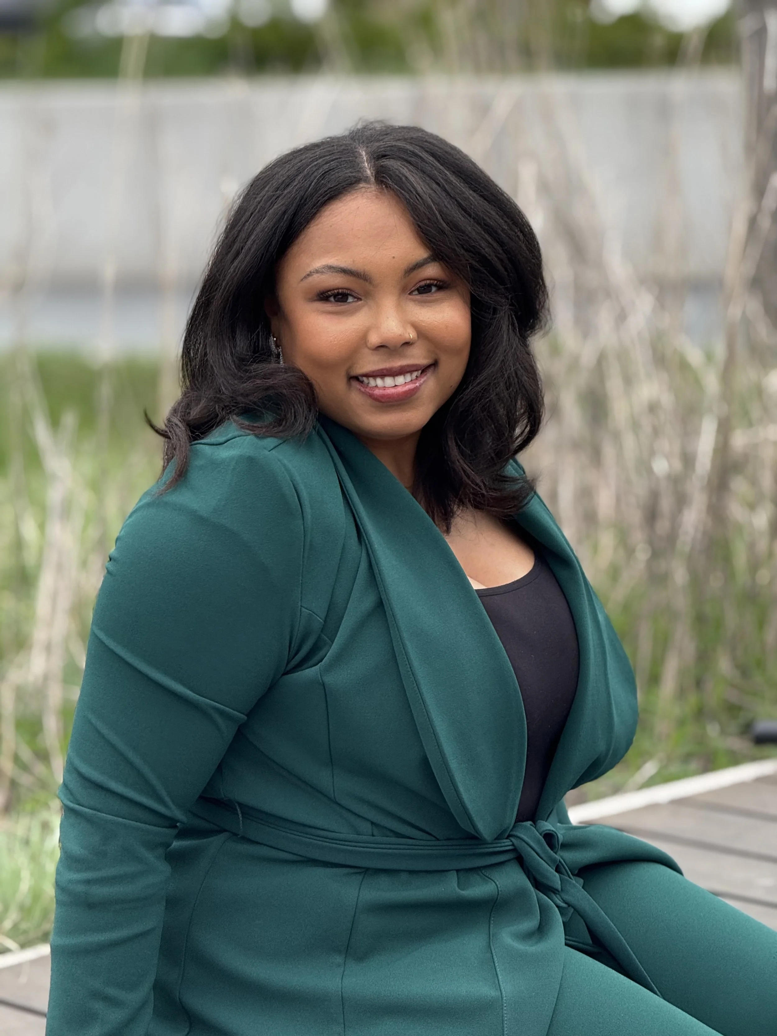 A woman with medium skin tone and long black hair sitting outside on a bench in a park, wearing a teal blazer and black top, smiling at the camera.