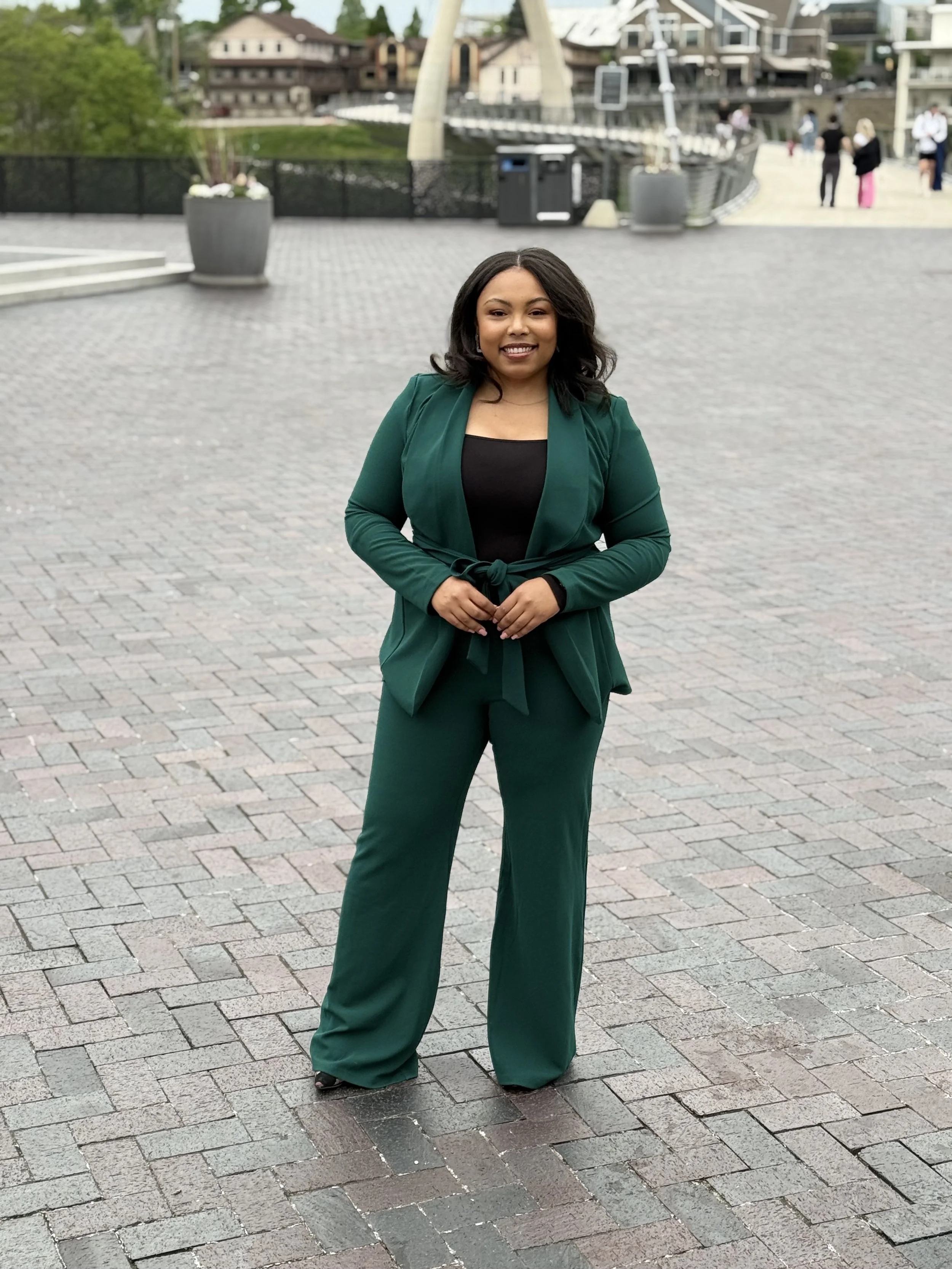 A woman with shoulder-length black hair, wearing a dark green suit with wide-leg pants and a matching blazer tied at the waist, standing outdoors on a paved area. She is smiling and has her hands clasped in front of her. In the background, there are buildings, people, large planters, and a bridge, indicating a scenic, urban setting.