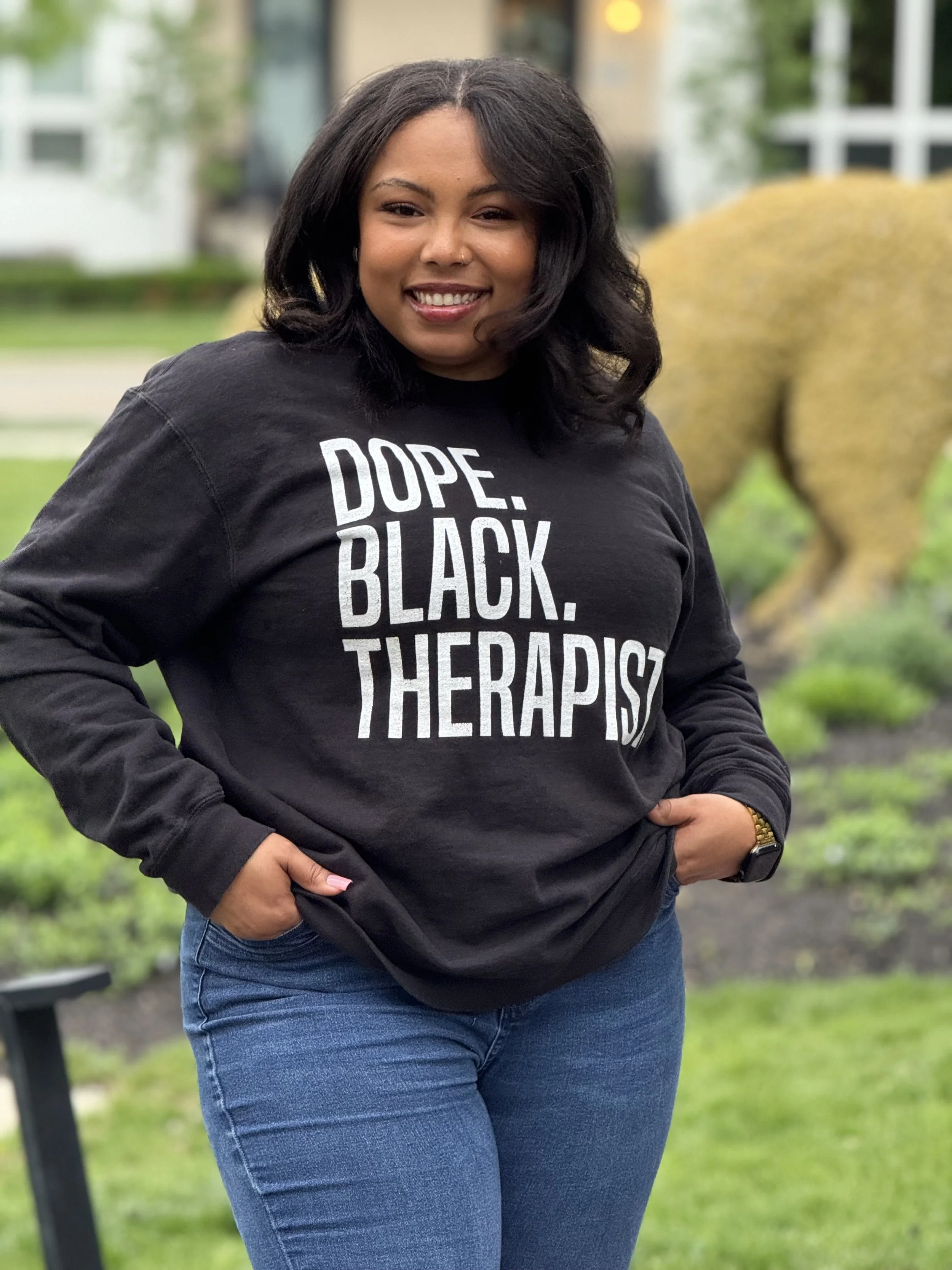 A woman standing outdoors with a smile, wearing a black sweatshirt with white text that says 'Dope. Black. Therapist', and blue jeans, with greenery and a yellow sculpture in the background.