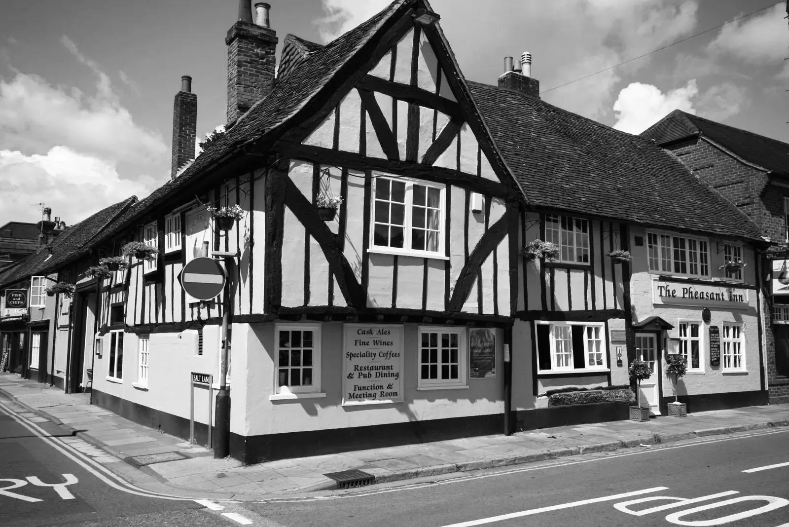 Black and white photo of a traditional English pub called "The Pheasant Inn," featuring timber-framed architecture, multiple windows with flower boxes, and signs advertising cask ales, fine wines, specialty coffees, restaurants, pub dining, functions, and a meeting room, located on a street corner.