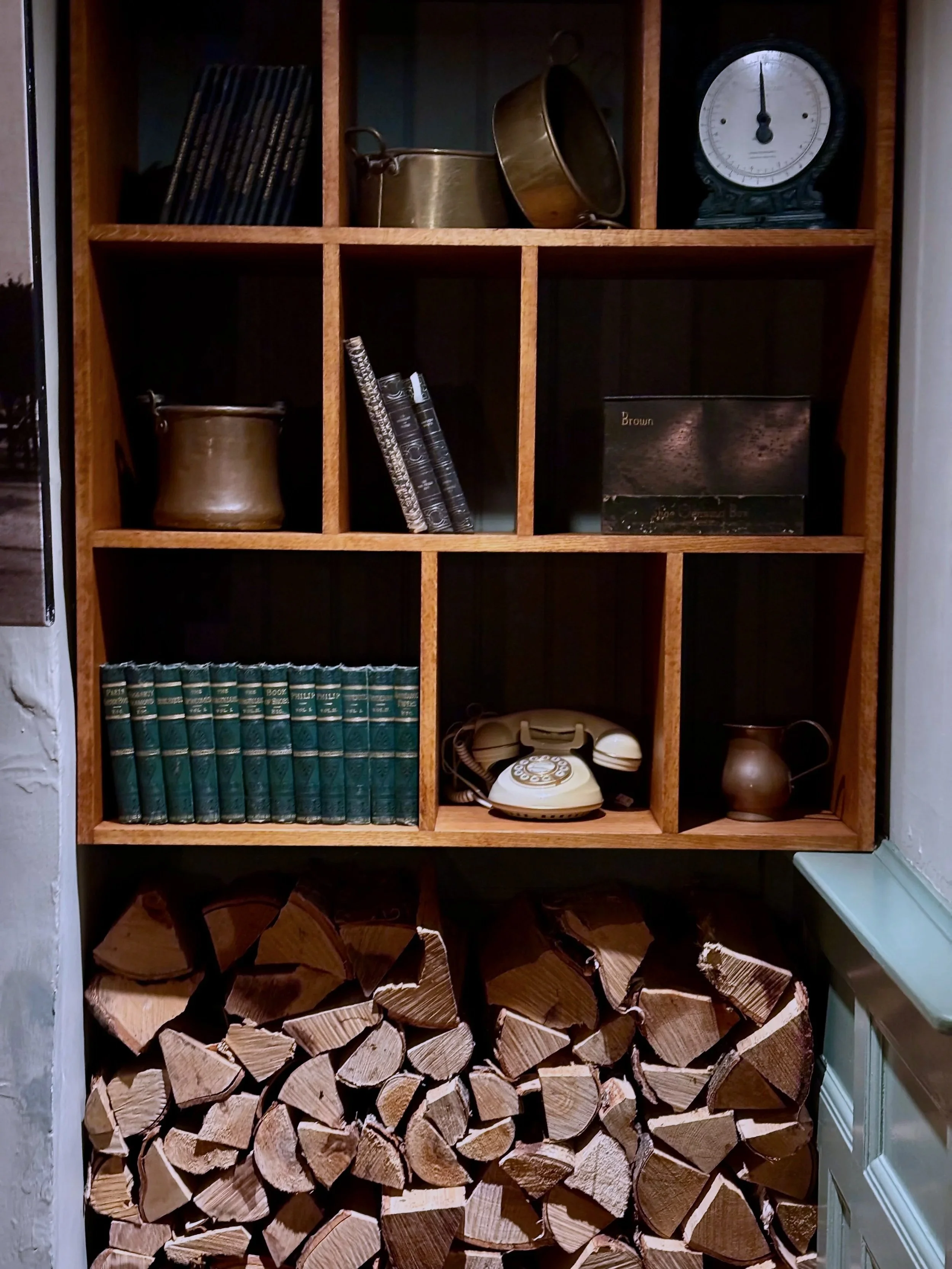Wooden shelves containing vintage books, brass pots, an old rotary telephone, a clock, and a collection of chopped firewood at the bottom.