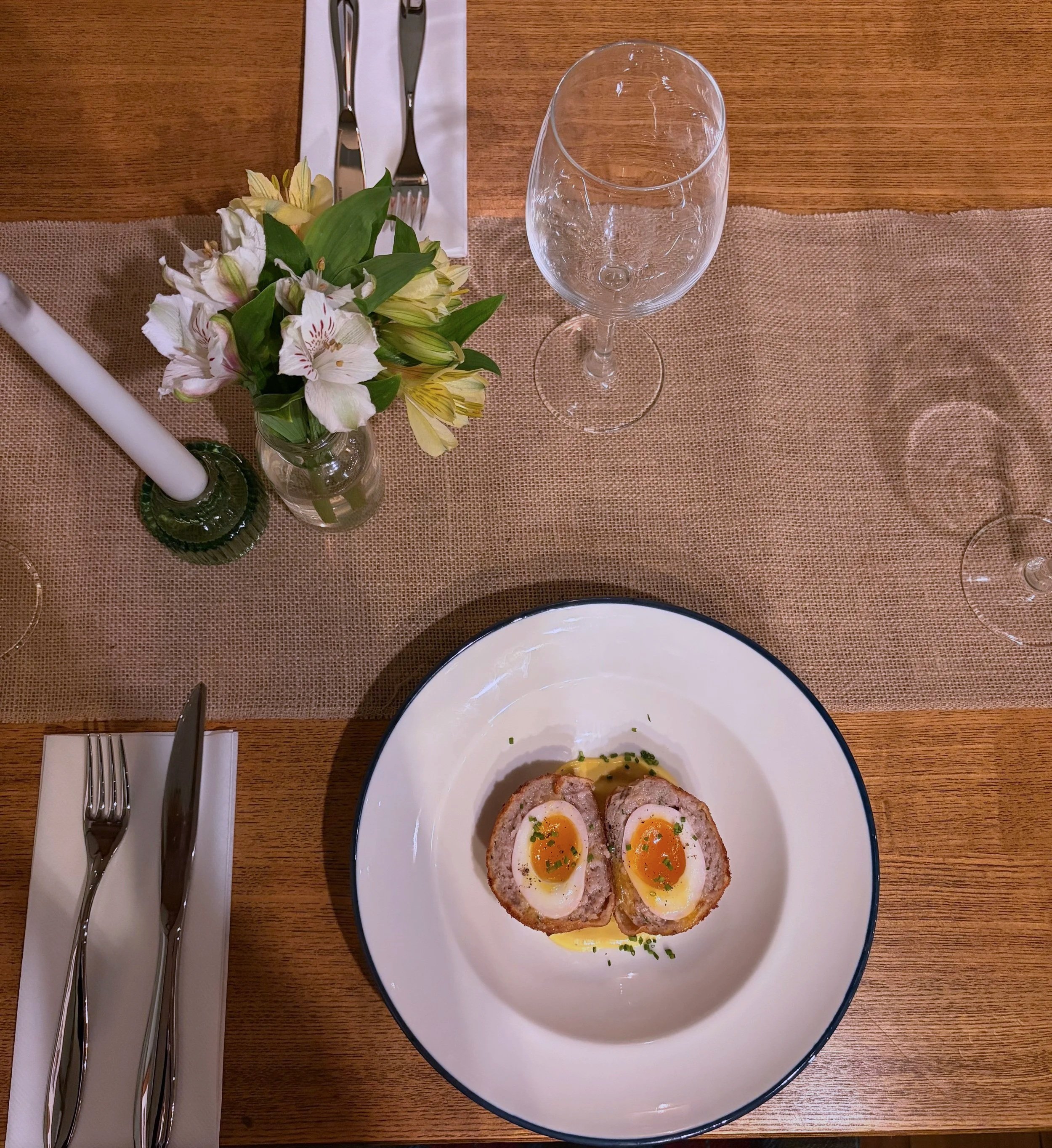 A table setting with a white plate containing halved Scotch eggs topped with soft-boiled egg yolks, chopped chives, and drizzled with sauce. The table has a brown burlap table runner, a small vase with white and yellow flowers, two empty wine glasses