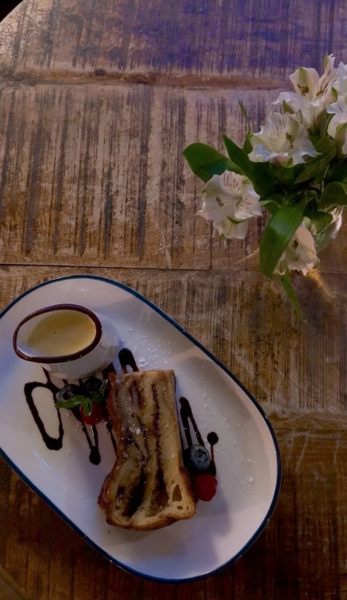 A dessert plate with a slice of French toast topped with powdered sugar, garnished with berries, a small cup of syrup, and drizzled with chocolate syrup, served on a white dish with a blue rim, alongside a glass vase of white flowers on a wooden tabl