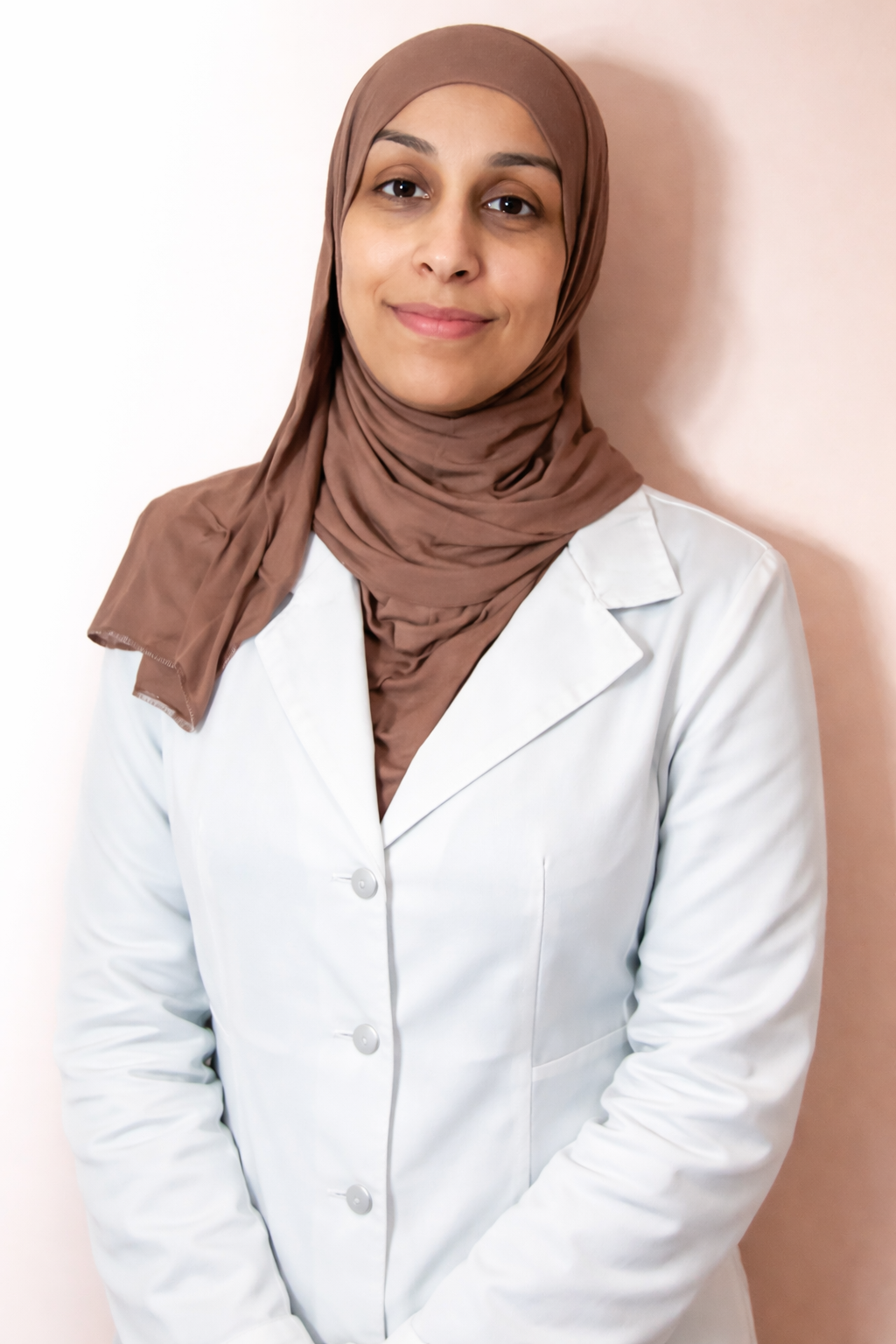 A woman wearing a brown hijab and a white lab coat, standing against a pink wall, smiling softly.