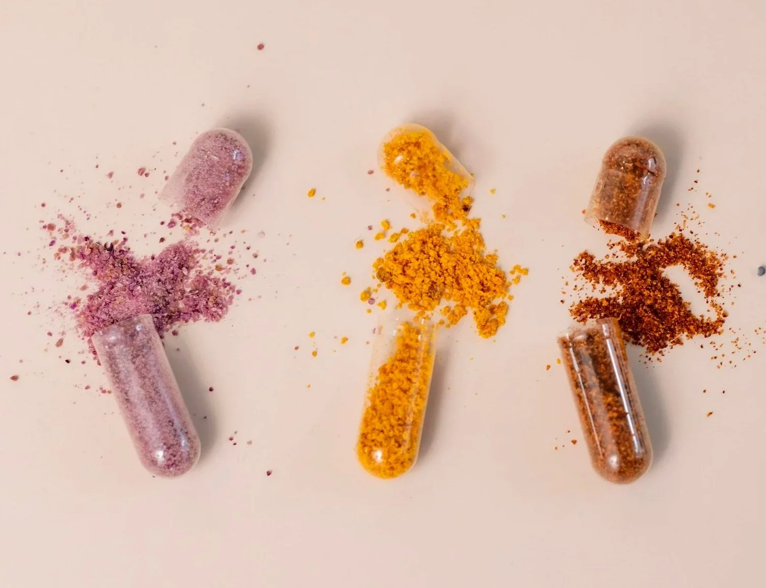 Three capsules with crushed powdered substances in pink, yellow, and brown colors on a white surface.
