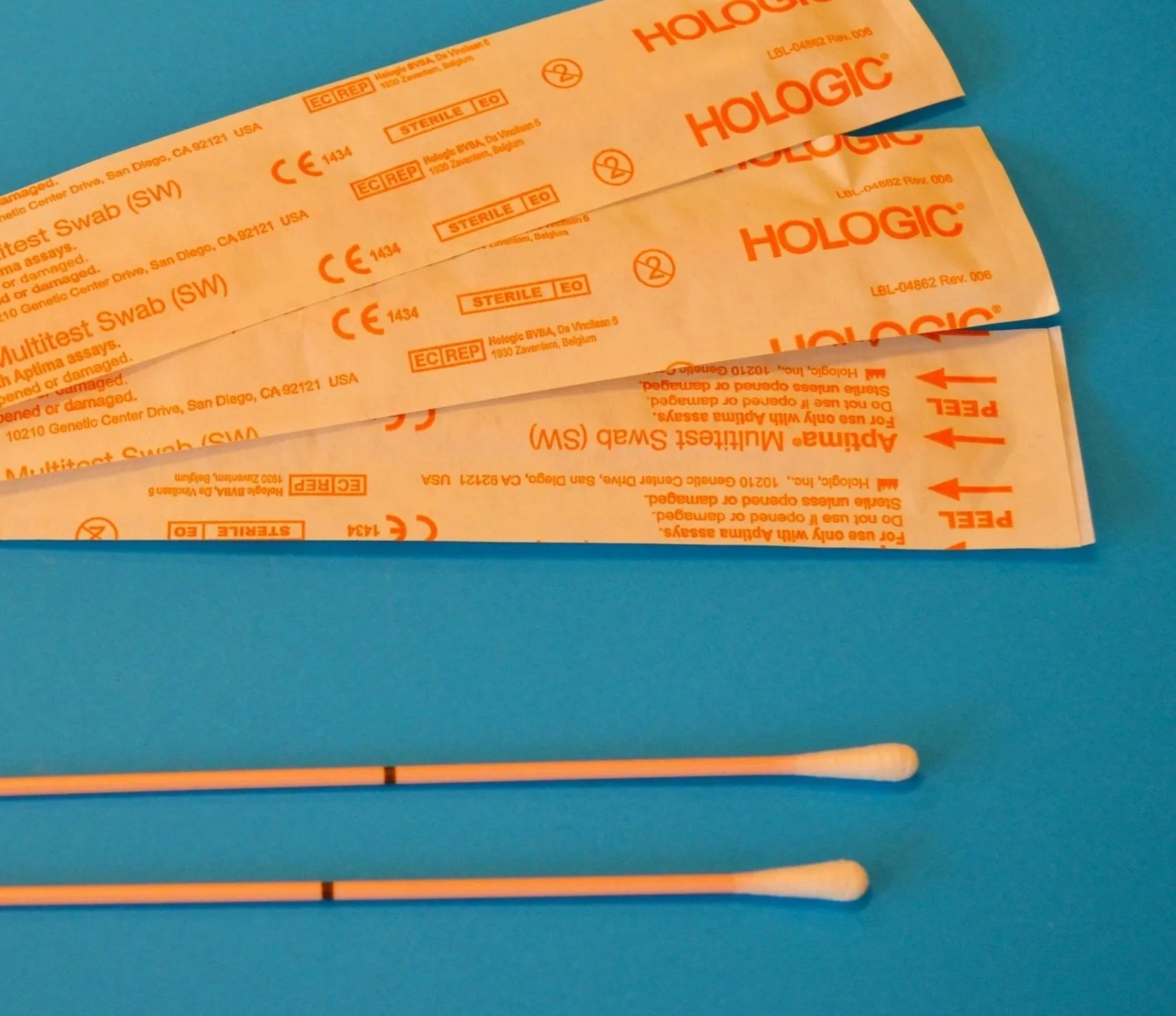Two sterile swabs and packaging for a Hologic multitest swab, with instructions and markings, lying on a blue background.
