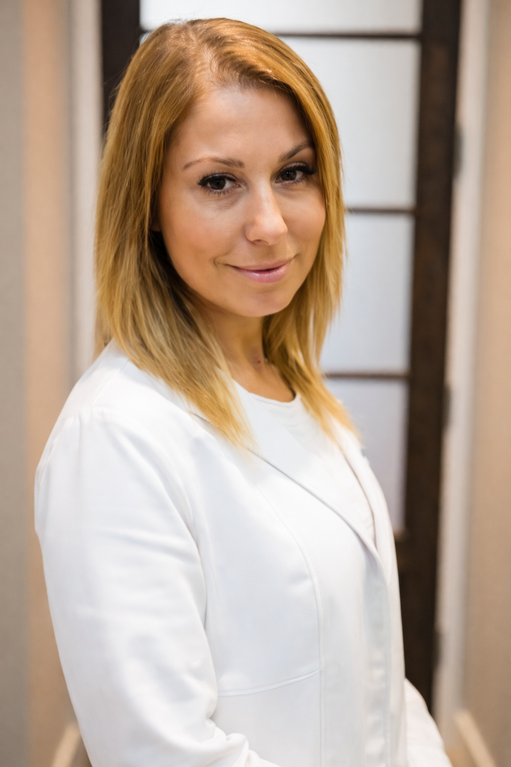 A woman with shoulder-length, reddish-blonde hair, wearing a white top, standing indoors beside a beige curtain and a wooden partition with frosted glass panels, smiling softly at the camera.