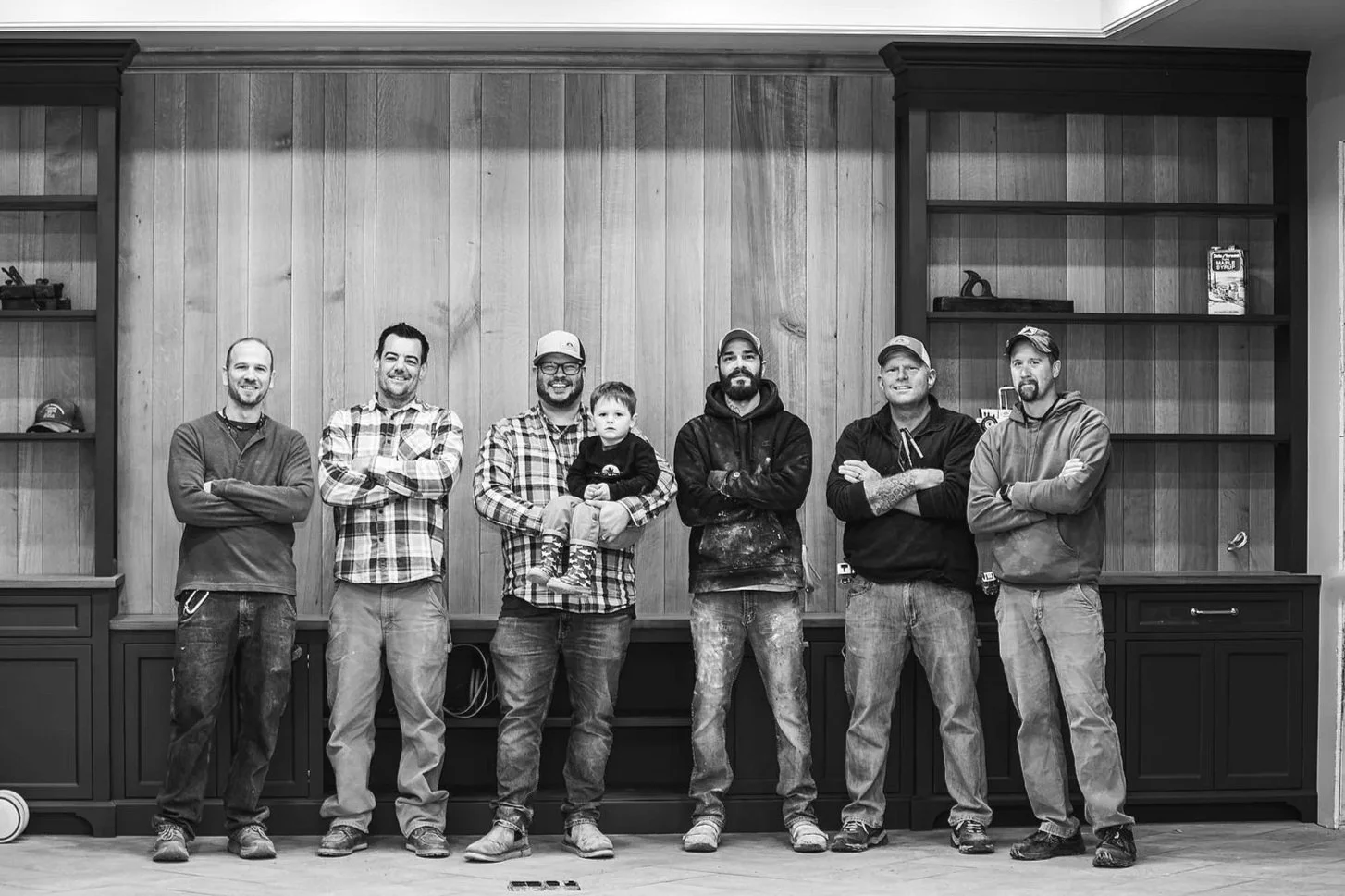 Seven men and one young boy standing in a row indoors, with a wood-paneled wall behind them and shelves on either side. The men are casually dressed, some with caps, and the boy is held by one man in the center. All are facing the camera with neutral or slight smiles.