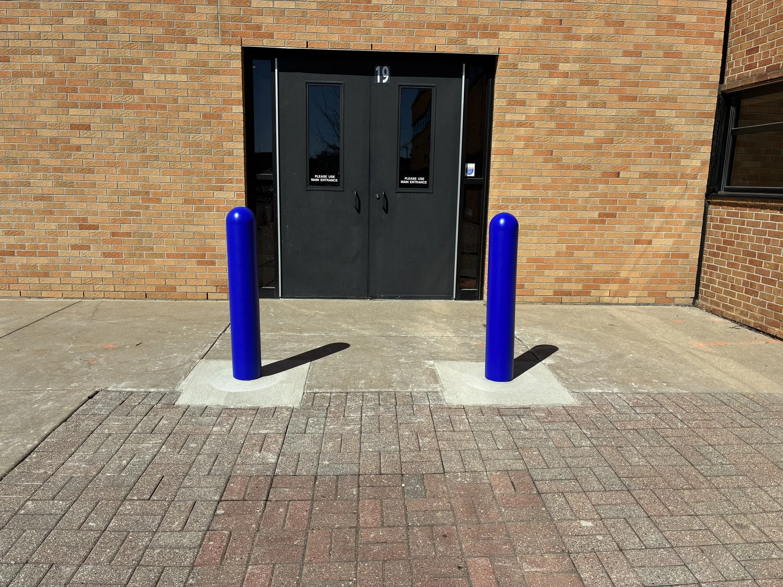 Two finished blue ASTM C40 crash-rated security bollards with decorative covers protecting side entrance door 19 at St. Mary Catholic School, Mount Clemens Michigan